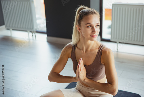 Portrait of young sportive happy girl, coach in sportswear doing yoga meditation on sports mat at meditation center, indoors. Concept of healthy lifestyle