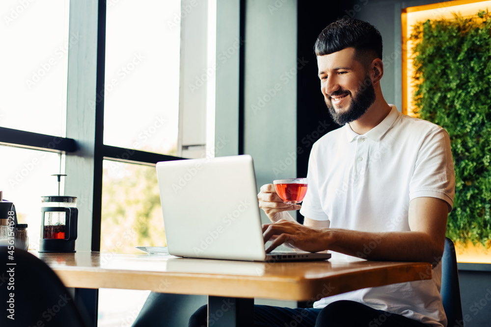 Smiling young using a laptop in a cafe and drinking tea while sitting at a wooden table, Male hands are typing on the laptop keyboard Smiling young using a laptop in a cafe and drinking tea while sitting at a wooden table, Male hands are typing on the laptop keyboard