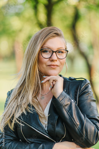 Young blonde woman in glasses for vision posing in the park in sunny weather on the body of a black jacket brutal image