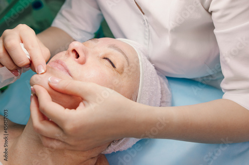 An elderly woman came to a beautician for a cleansing procedure. The hands of a specialist do massage and facial cleansing