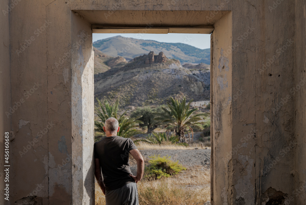 Rear view of adult man standing in doorway of an abandoned house looking at view