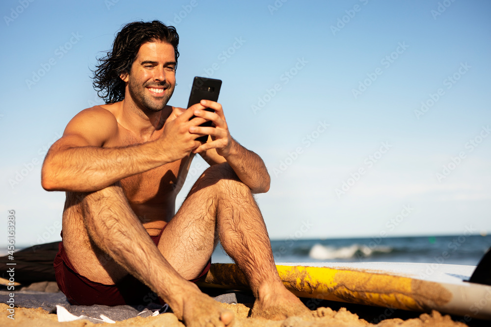 Portrait of handsome surfer with his surfboard. Young man using the phone while relaxing on the beach.