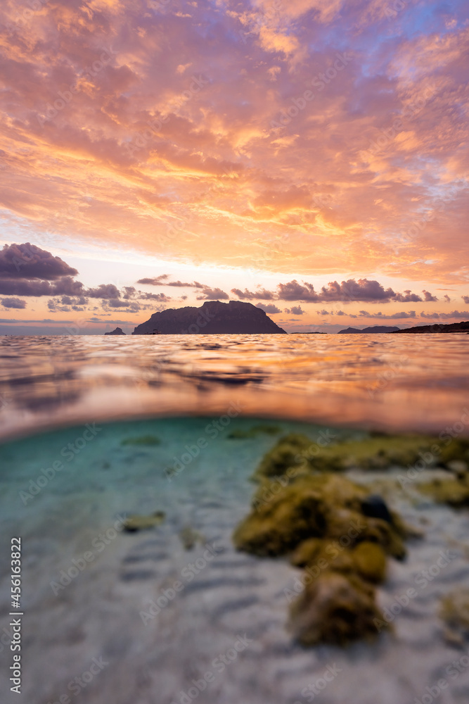 (Selective focus) Split shot, over under water surface. Defocused waves in the foreground with Tavolara Island on the surface during a dramatic sunrise. Porto Istana, Sardinia, Italy.