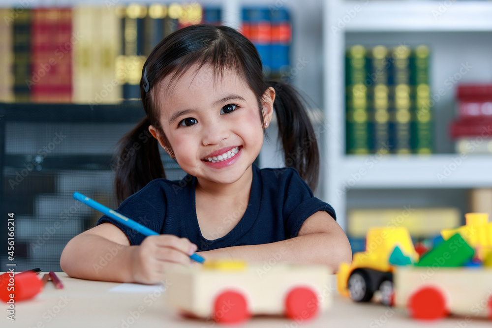 Small pretty pigtails hair preschooler kindergarten happy girl sit on chair drawing with colored pencils on paper on table full of plastic truck toys in living room at home in front bookshelf Small pretty pigtails hair preschooler kindergarten happy girl sit on chair drawing with colored pencils on paper on table full of plastic truck toys in living room at home in front bookshelf