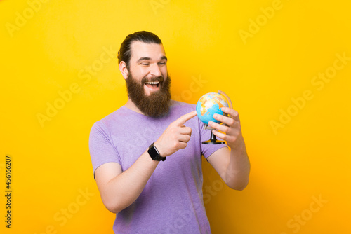 Smiling bearded man is pointing on a small globe over yellow background. photo