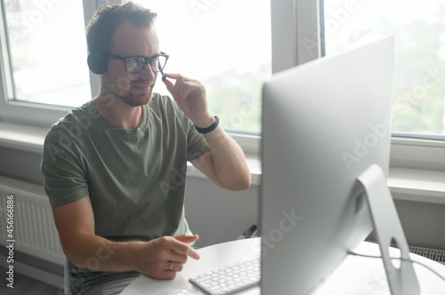 A confident male freelancer sitting at the desk with a laptop, using headset communicating with customers, a helpful support officer speaking online and makes marks on computer app, side view