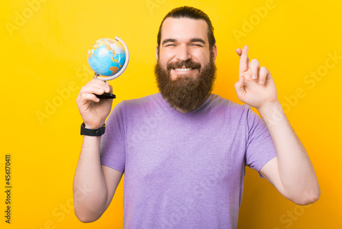 Young bearded man is holding a small globe and crossing fingers over yellow background. photo