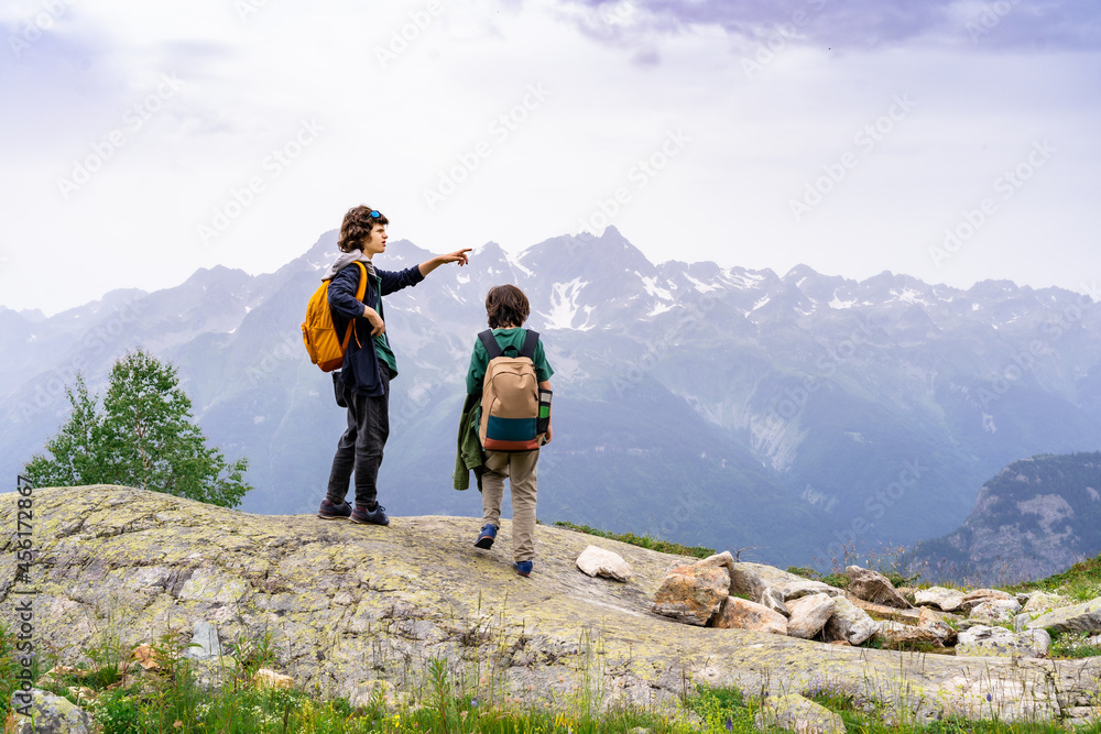 Two boys with backpacks behind them standing on the top of the mountain, resting after a long ascent. Elder boy pointing into the distance. Active vacation concept. Hiking in family.