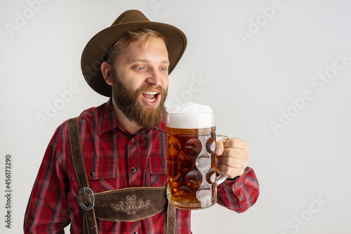 Half-length portrait of bearded man in hat and traditional Bavarian costume holding huge mug, glass of light frothy beer isolated over white background. Flyer