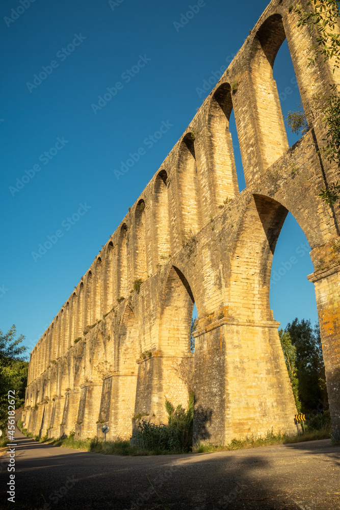 Vertical photograph of the Pegões Aqueduct, near the city of Tomar in Portugal, lit by the sun at the end of the day with a blue sky in the background.