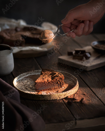 Chocolate cake on a wooden table. A piece of brownie on a brown background. Cocoa and chocolate pieces. Serving dessert. A man's hand pours cocoa on a cupcake