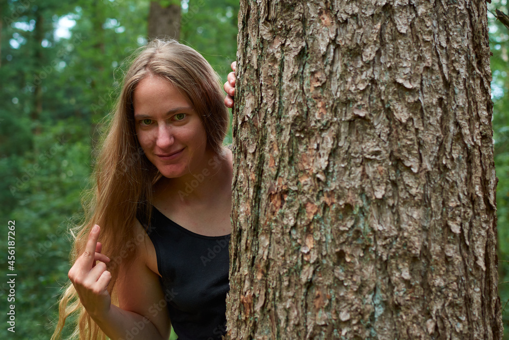 A blonde Caucasian lady with long hair looking from behind a tree trunk showing a middle finger A blonde Caucasian lady with long hair looking from behind a tree trunk showing a middle finger