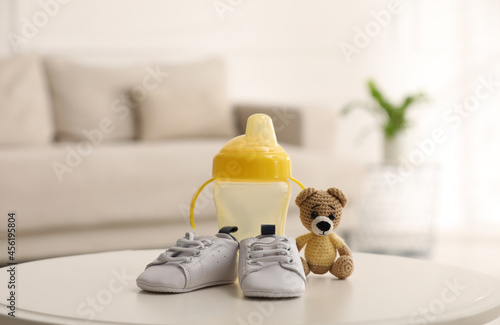 Baby bottle and shoes near toy bear on white table in room. Maternity leave concept