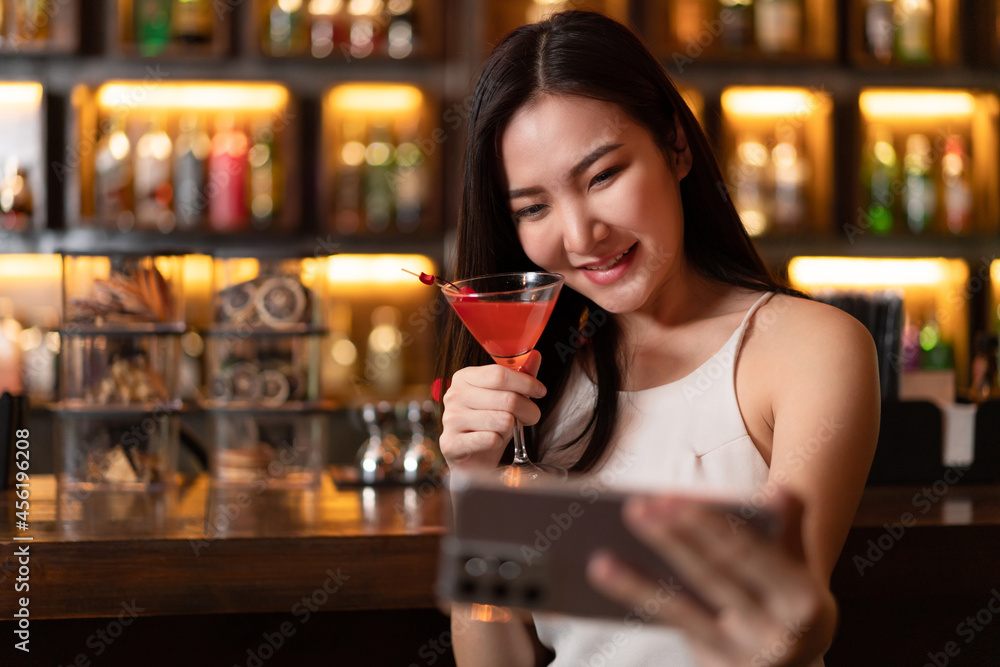 Nightlife concept a sexy girl with smiling face doing selfie on her smartphone while hold a vivid pink champaign in the night bar Nightlife concept a sexy girl with smiling face doing selfie on her smartphone while hold a vivid pink champaign in the night bar