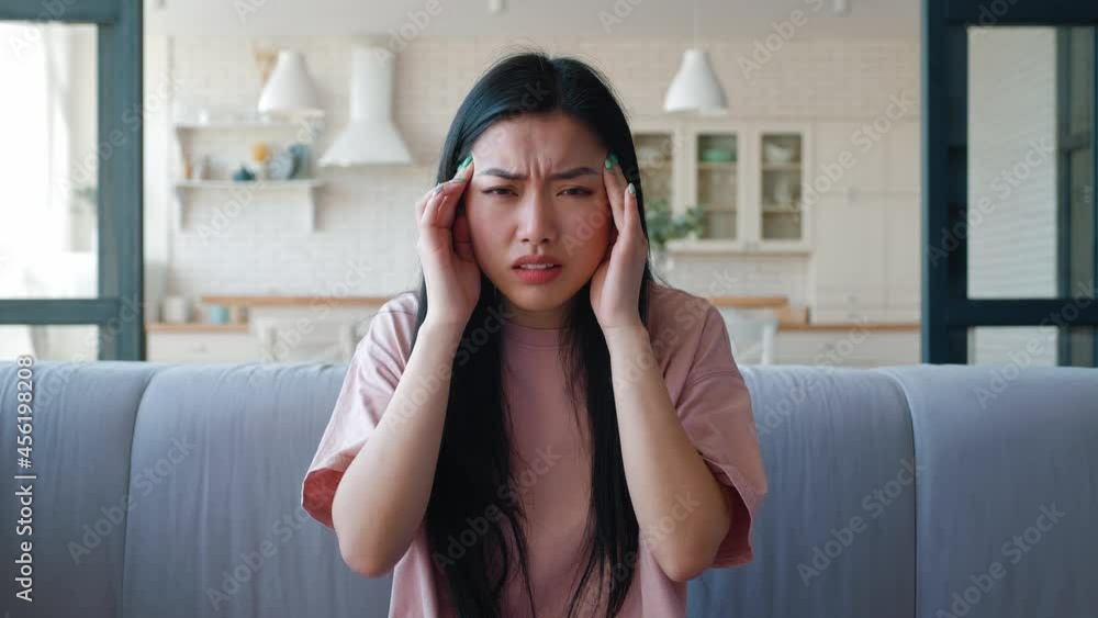 Frustrated deappointed and upset young Asian woman sitting on the couch in living room looking at camera, feeling chock, massaging her temple. Main focus on facial expressions and negative emotions