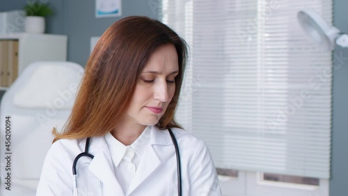 Adult caucasian woman in white medical gown sitting at her workplace in hospital. Cheerful female doctor with stethoscope looking to the camera and smiling. Medicine concept photo