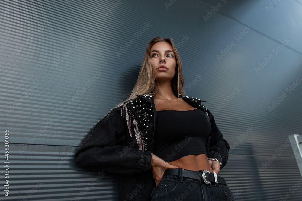 Beautiful stylish girl in a cool fashionable black denim jacket with black jeans and a t-shirt posing near a modern metal wall in the city Beautiful stylish girl in a cool fashionable black denim jacket with black jeans and a t-shirt posing near a modern metal wall in the city