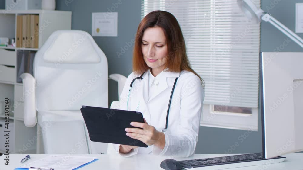 Competent caucasian female doctor with stethoscope working on tablet computer in hospital office. Adult woman in white gown making medical reports. Profession and work concept Competent caucasian female doctor with stethoscope working on tablet computer in hospital office. Adult woman in white gown making medical reports. Profession and work concept