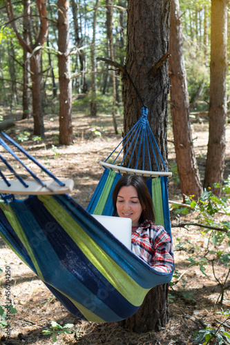 Freelance and working at nature. Smiling woman freelancer works at notebook while lying on the hammock in the forest. Online business independent of the office.