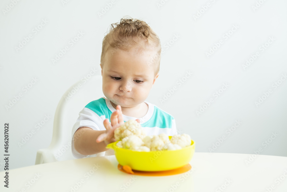 smiling baby boy sitting in a Childs chair eating cauliflower on white kitchen. smiling baby boy sitting in a Childs chair eating cauliflower on white kitchen.