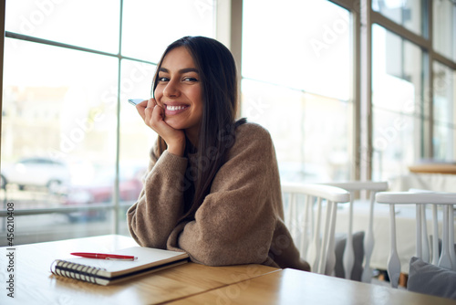 Portrait of cheerful Middle Eastern female student with education notepad posing at table desktop in university campus and smiling at camera during learning break, happy South ASian blogger in cafe