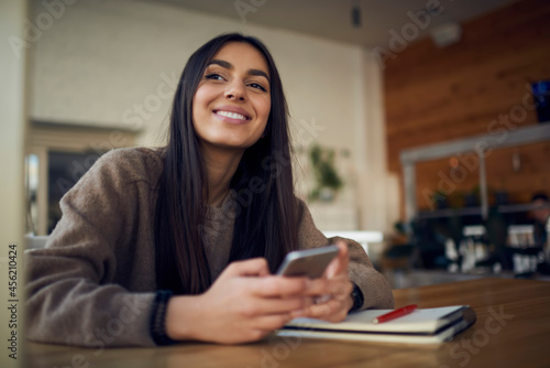 Happy female blogger with smartphone and personal planner for creating content ideas looking away and smiling during school time for learning at desktop  cheerful millennial woman with cellphone