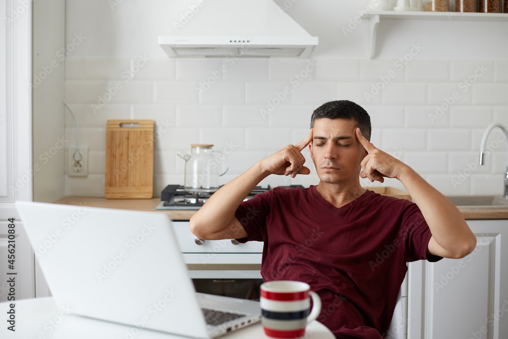 Indoor shot of exhausted man posing in kitchen while sitting at table in front laptop, feels tired, keeps eyes closed, massaging his temples with fingers, suffering from headache.
