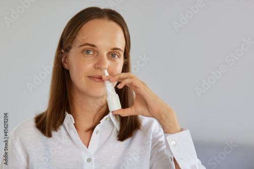 Closeup indoor shot of charming woman using nasal spray for runny nose, catches cold, looking at camera, wearing white shirt, posing against light wall.