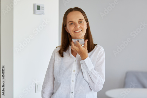 Indoor shot of satisfied young adult woman with dark hair holding smart phone in hands, recording voice message, looking at camera with positive facial expression.