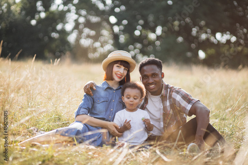 African american father and caucasian mother enjoying summer picnic with their cute little son. Beautiful family sitting on grass and smiling sincerely. Relaxation and love concept.