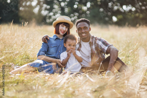Positive multicultural parents sitting with little son on grass and enjoying summer picnic on field. Caucasian mother and african father embracing their lovely child.