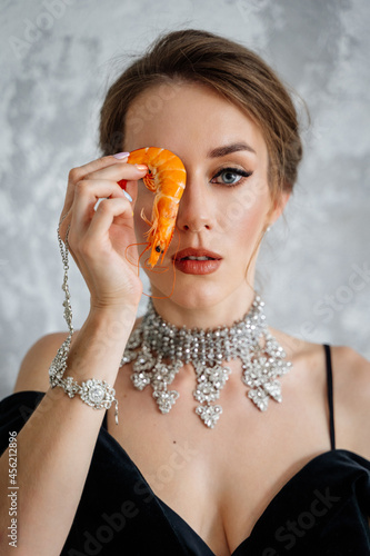 beautiful young woman in a swimsuit sitting on the floor near the window and looking away holds shrimp