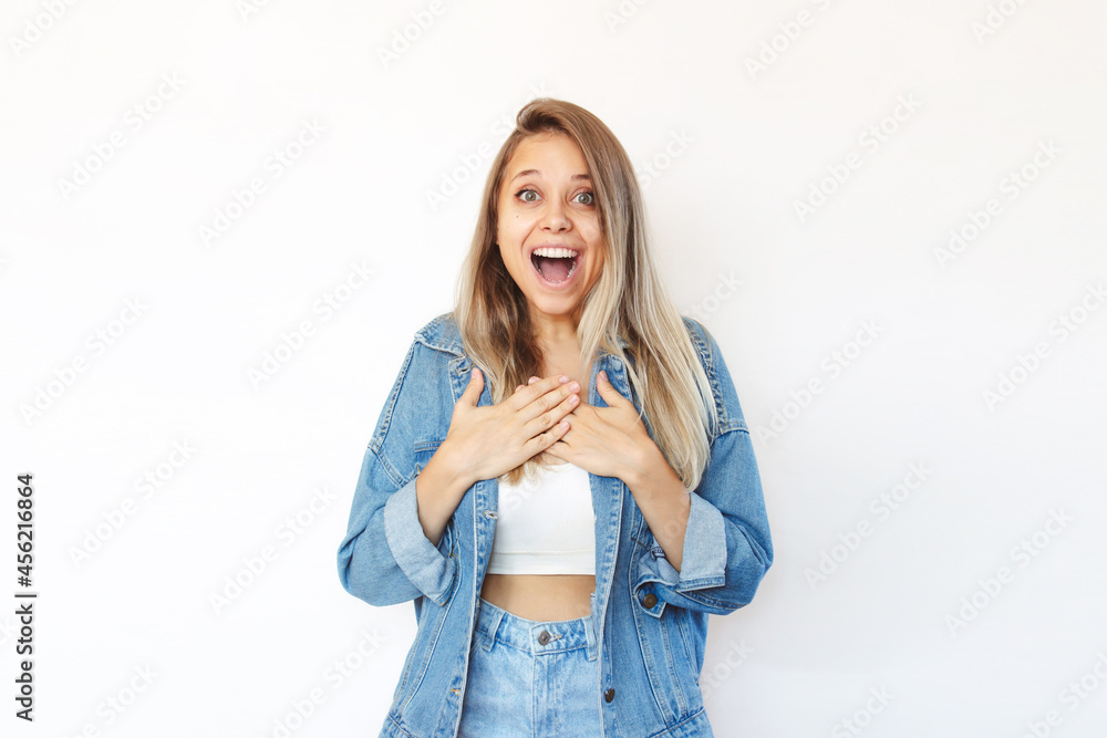 A young caucasian smiling amazed happy blonde woman in a denim jacket and jeans holding her hands on a chest is surprised and glad to hear the good news about something isolated on a white background A young caucasian smiling amazed happy blonde woman in a denim jacket and jeans holding her hands on a chest is surprised and glad to hear the good news about something isolated on a white background