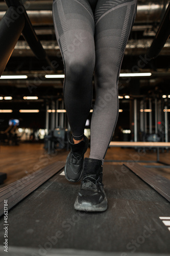Beautiful women's athletic legs in black sportswear with black sneakers running on the treadmill at the gym