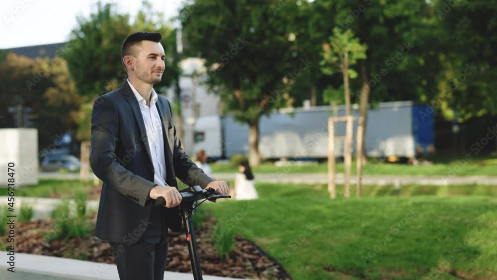 Slow-motion side-view of caucasian businessman riding on electric scooter along sidewalk on warm sunny day. Modern transportation gadget and popular futuristic device among young people. Slow-motion side-view of caucasian businessman riding on electric scooter along sidewalk on warm sunny day. Modern transportation gadget and popular futuristic device among young people.