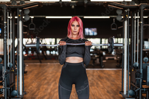 Beautiful fitness model woman in black stylish sportswear workout and trains with a barbell in the gym and looks at the camera