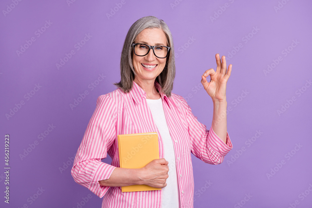 Photo of mature woman happy smile show okay alright ad sign hold book materials isolated over purple color background Photo of mature woman happy smile show okay alright ad sign hold book materials isolated over purple color background