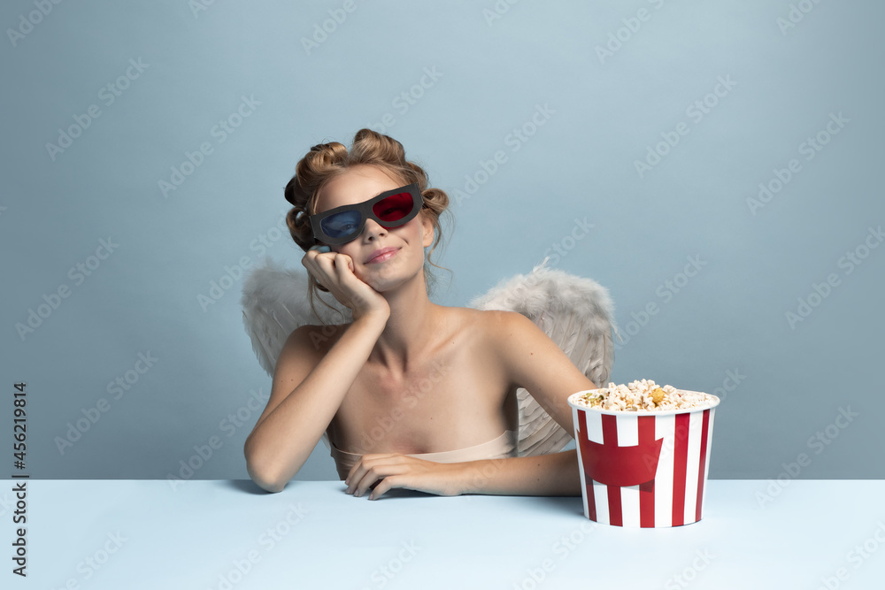 Obraz premium Portrait of young beautiful girl in image of angel with wings sitting and watcjing film isolated on blue gray studio background. Concept of beauty, purity