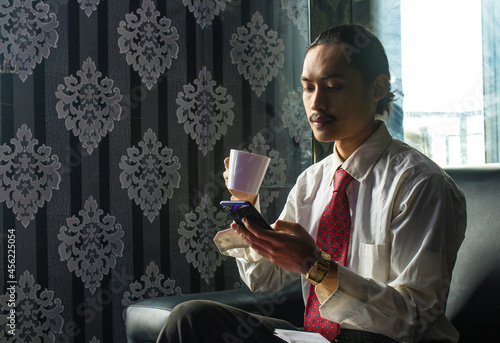 Relaxed Smilling Asian Man drinking coffee while using a cell phone for work