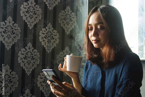 Relaxed beautiful Asian Woman drinking coffee while using a cell phone for work