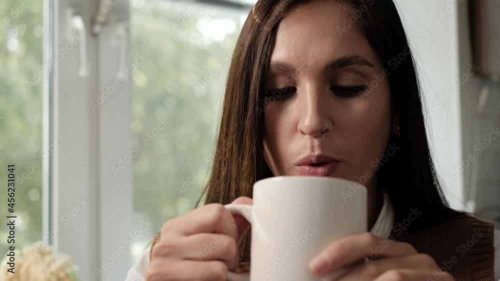 Woman drinking coffee and enjoys taste. Face of happy woman with cup of coffee or tea in her hand, she drinks drink and looks out window. Close-up and slow motion Woman drinking coffee and enjoys taste. Face of happy woman with cup of coffee or tea in her hand, she drinks drink and looks out window. Close-up and slow motion