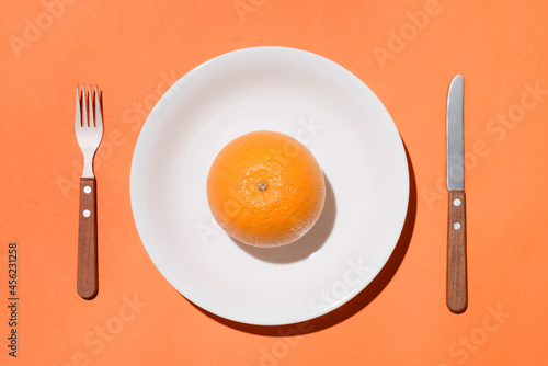 Top view of orange fruit on white plate with knife and fork on orange background