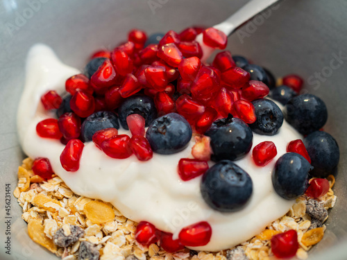 a light gray bowl with a spoon. In the bowl there are oatmeal, muesli, yogurt and various types of fruit, pomegranate, blueberries. In close-up