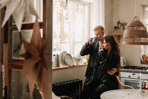 Married man and woman in love in pajamas having breakfast drinking tea sitting at the dining table in the kitchen in an eco style interior during the winter holidays in a cozy country house