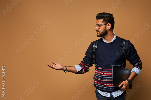 Stylish handsome indian student with laptop and backpack pointiong his hand to background photo