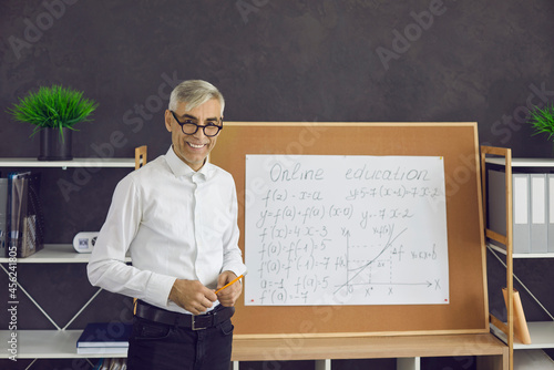 Portrait of a friendly senior male teacher who remotely conducts a math lesson for his students. Man stands at the blackboard and explains mathematical formulas online. Online education concept.