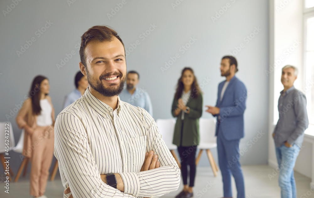 Close up portrait of a successful young business man who is looking at camera with confident expression. Man stands against the background of people talking to each other in a bright hall.