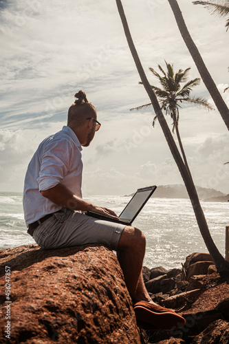 Cyfrowy nomada, człowiek pracujący z laptopem i smartfonem zdalnie na tle oceanu i skał siedzący na wybrzeżu.