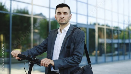 Portrait shot of caucasian man leaning on electric scooter and looking at camera. Stylysh man on vehicle outdoors. Eco-friendly modern urban transport photo