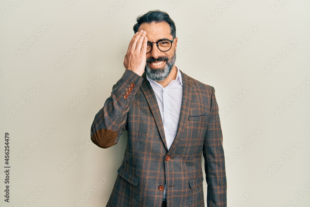 Middle age man with beard and grey hair wearing business jacket and glasses covering one eye with hand, confident smile on face and surprise emotion. Middle age man with beard and grey hair wearing business jacket and glasses covering one eye with hand, confident smile on face and surprise emotion.